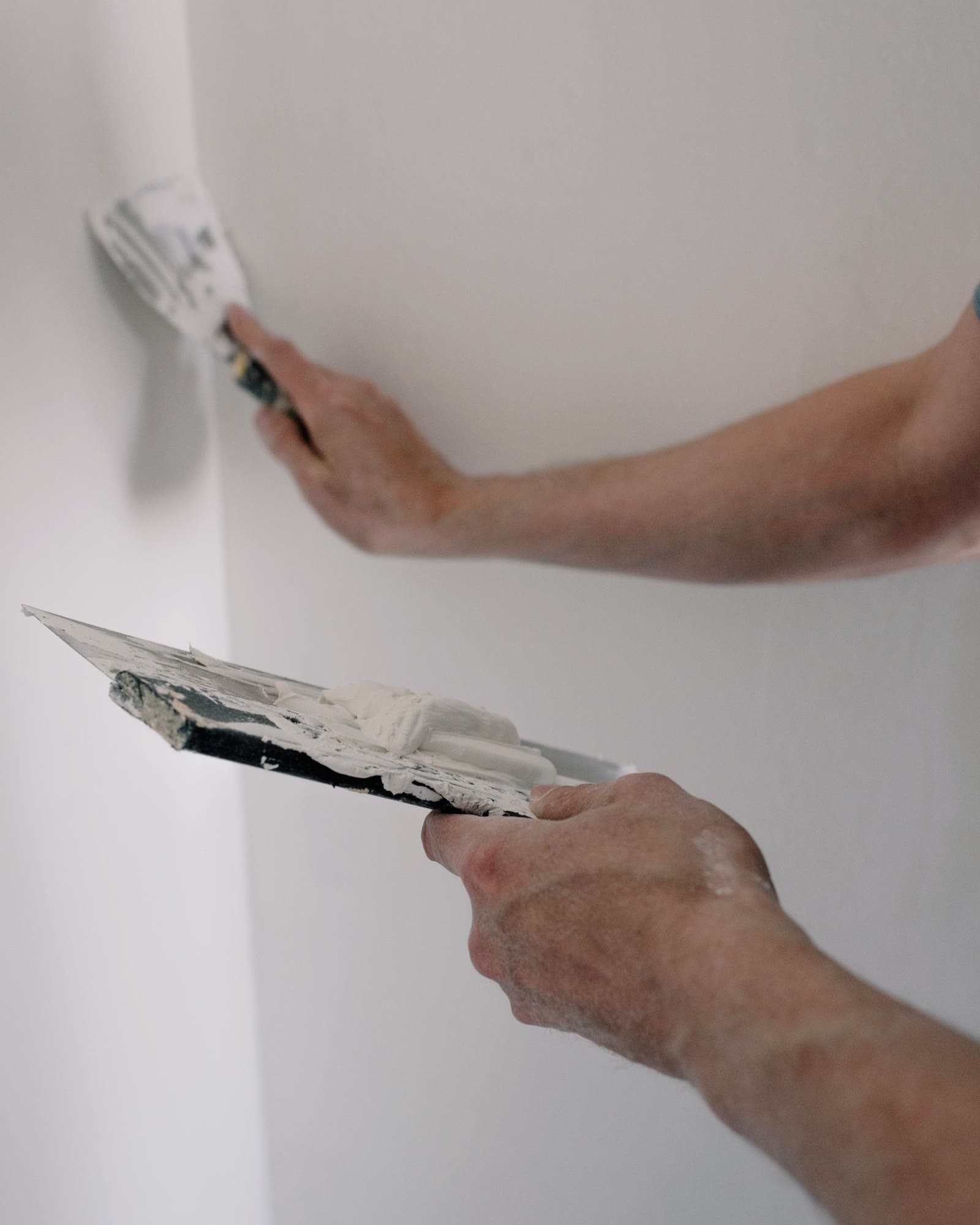 Hands working joint compound into a wall corner with two putty knives